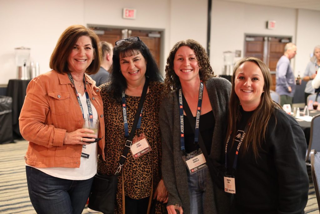 Four women stand together indoors at the Accelerate Event in Bloomington MN, smiling at the camera with ASB lanyards and name badges around their necks. Other people and tables are visible in the background.