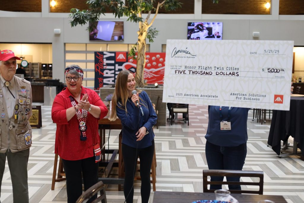 Four people stand indoors, smiling, as one woman holds a giant $5,000 check from ASB made out to Honor Flight Twin Cities. The setting appears to be a public event or donation ceremony, possibly part of an Accelerate Event.