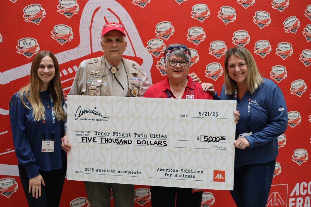 Four people stand in front of a red backdrop, smiling and holding an oversized $5,000 check made out to Honor Flight Twin Cities from ASB at the Accelerate Event, dated 5/21/25.
