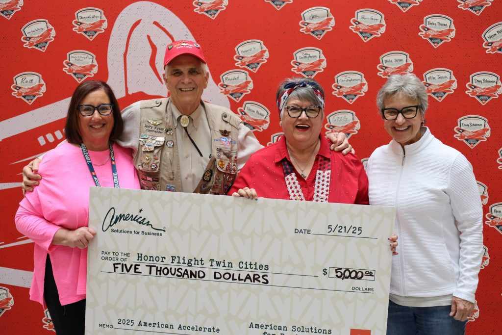 Four smiling adults hold a $5,000 ceremonial check for Honor Flight Twin Cities at the ASB Accelerate Event in Bloomington MN. One person wears a decorated uniform; the others are in casual clothing, standing before a red logo backdrop.