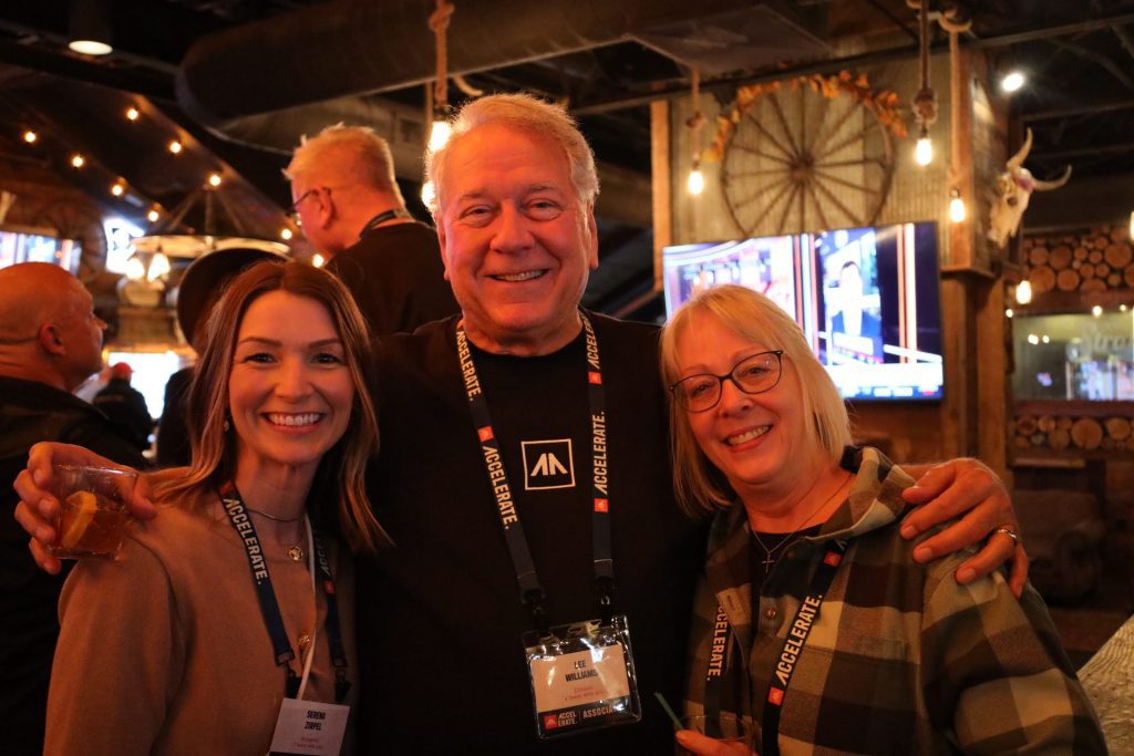 Three people smile together at the ASB Accelerate Event in Bloomington MN, wearing lanyards and name badges. The man in the center has his arms around the two women. The background features warm lighting and rustic decor.
