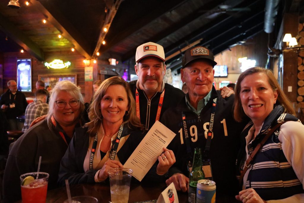 Five people smiling and posing together at a bar during the ASB Accelerate Event in Bloomington MN, some holding drinks and a menu. They wear lanyards and casual jackets, with warm lighting and a rustic atmosphere in the background.