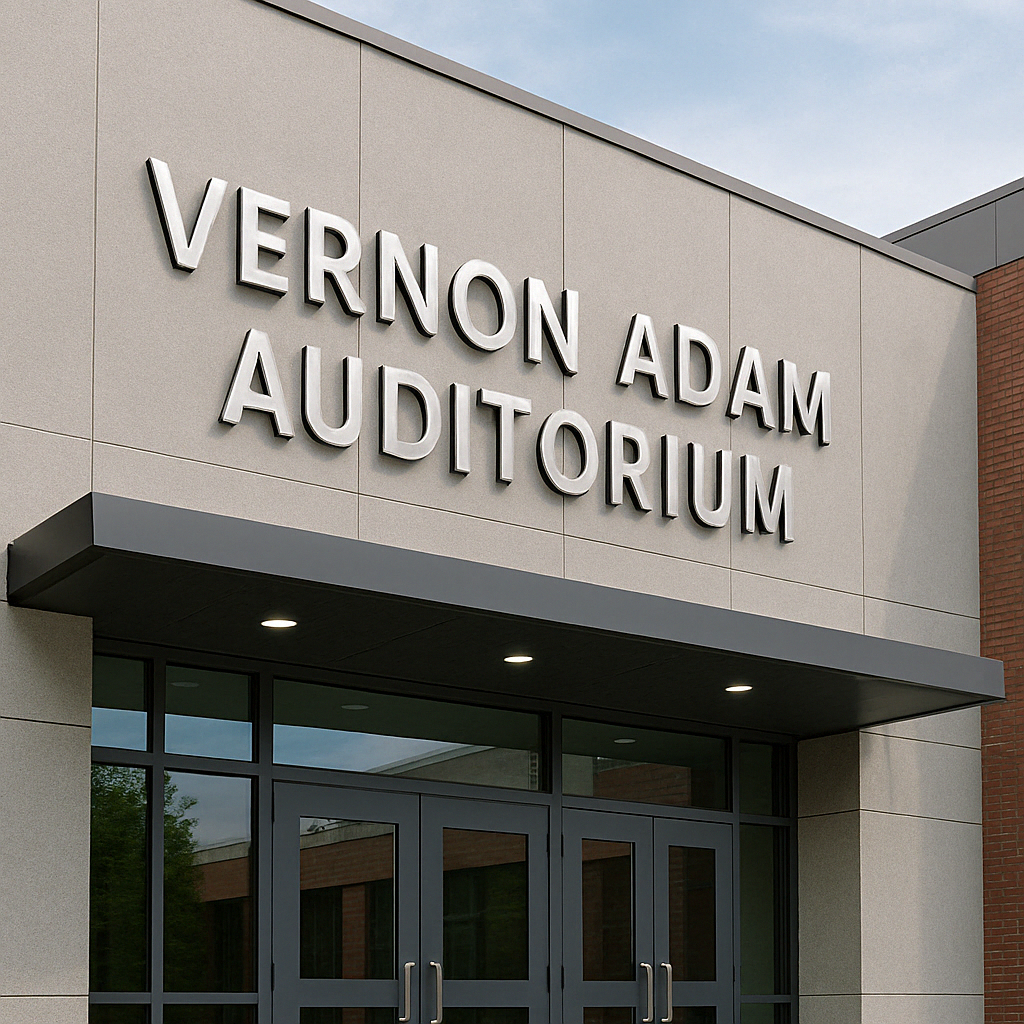 The entrance of a building labeled "Vernon Adam Auditorium" in large, metallic letters above double glass doors showcases striking signage solutions, with part of a brick wall and a cloudy sky visible.