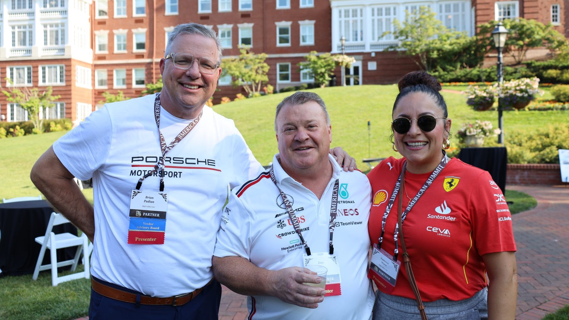 Spotlight: Three people stand smiling outdoors in front of a large brick building. Two men wear motorsport shirts and lanyards, while a woman in a red Ferrari shirt and sunglasses enjoys the scene—green lawns and white chairs complete the Vendor Advisory event.
