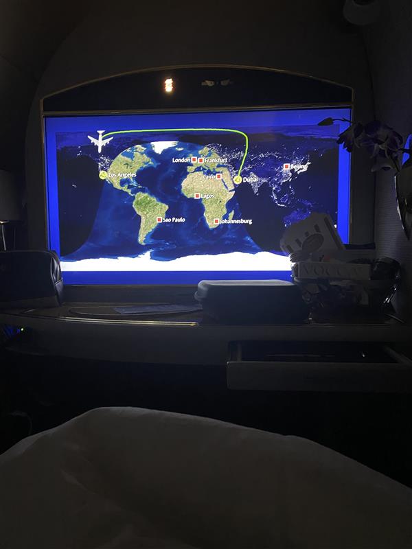 In-flight display spotlight showing a world map with a highlighted flight path from Dubai to New York, visible from a passenger’s seat with part of a blanket and cabin amenities in the foreground.