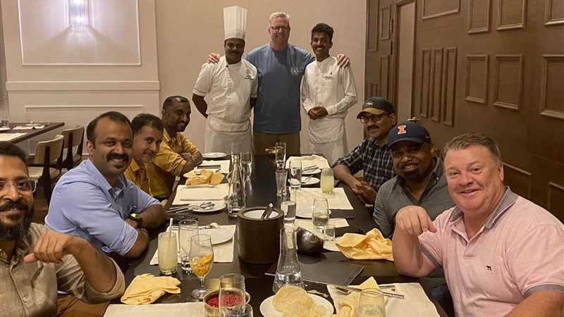 Nine men, including two chefs in white uniforms, pose and smile around a dining table set with drinks and food in a warmly lit restaurant for a Vendor Spotlight celebration with Brian Padian. The group appears to be enjoying a special gathering.