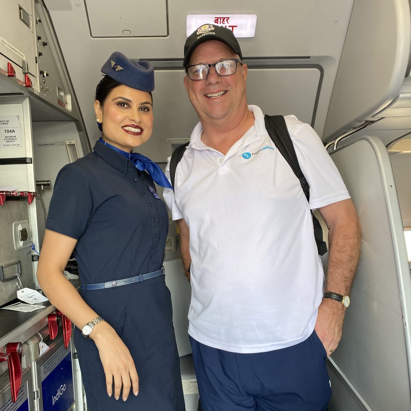 A smiling flight attendant in a navy uniform stands next to Brian Padian, who is in a white polo, navy shorts, and black cap, inside an airplane near the doors and galley—truly a Spotlight moment.