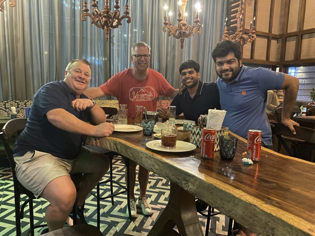 Four men sit around a wooden table in a restaurant, smiling at the camera. Drinks, plates, Coca-Cola cans, and glasses are on the table. Decorative chandeliers hang above and patterned tiles cover the floor, putting a spotlight on this cheerful gathering.