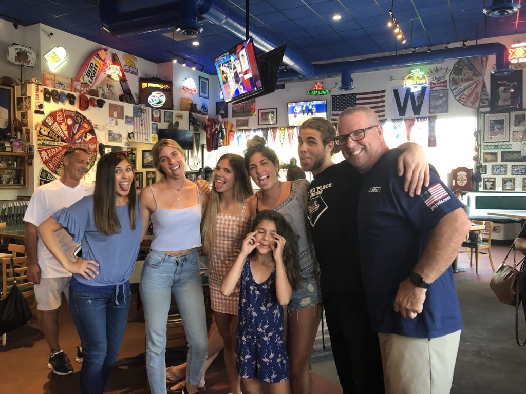 A group of seven people, including a young girl, pose playfully and smile inside Brian Padian’s lively, colorful restaurant filled with wall decorations and sports memorabilia—a perfect spot for Vendor Advisory gatherings.