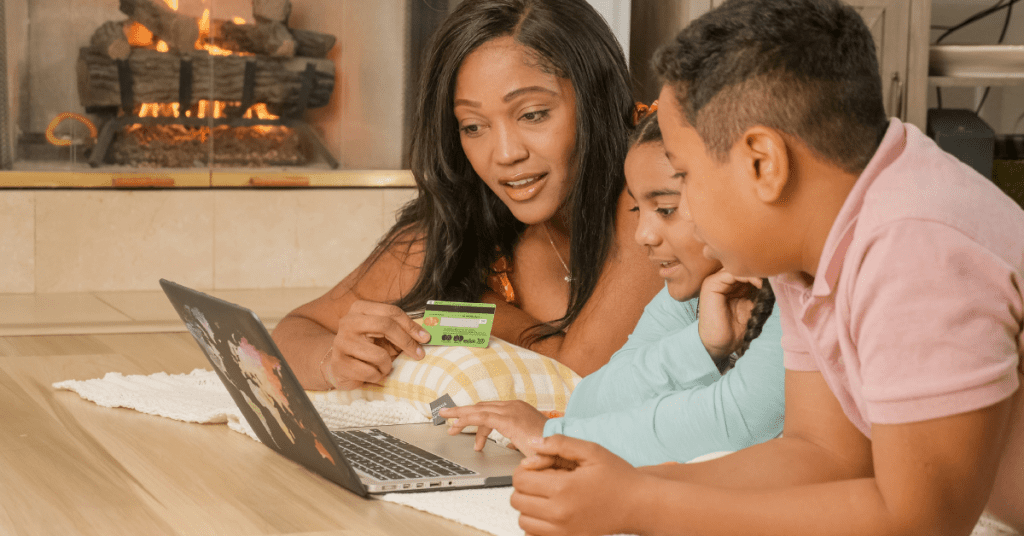 A woman and two children lie on the floor in front of a fireplace, looking at a laptop. The woman holds a green credit card, suggesting they’re exploring Youth Accounts from Banks and building lasting relationships together.