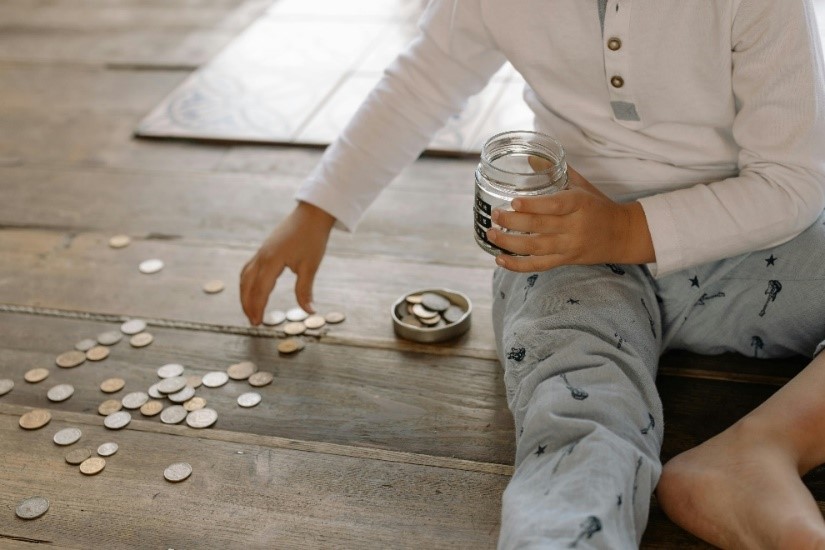 A child sits on a wooden floor, holding a jar of coins and reaching for scattered coins, as more pile up in a small dish nearby—an early lesson in saving that banks nurture with youth accounts for lasting relationships.