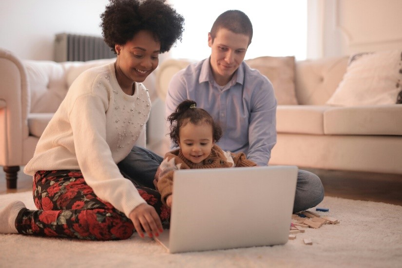 A smiling family of three sits on the living room floor, happily using a laptop together. The child is in the center with the adults on either side, all focused on the screen—building lasting relationships while exploring youth accounts from banks.