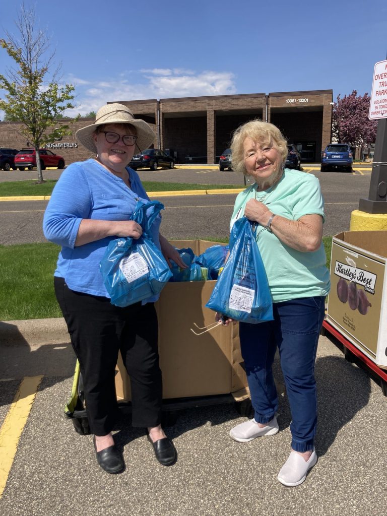 Two smiling women stand outdoors next to a large box, each holding blue plastic bags filled with items during the American Solutions for Business annual food drive. It's a sunny day, with buildings, cars, and a grassy area in the background.