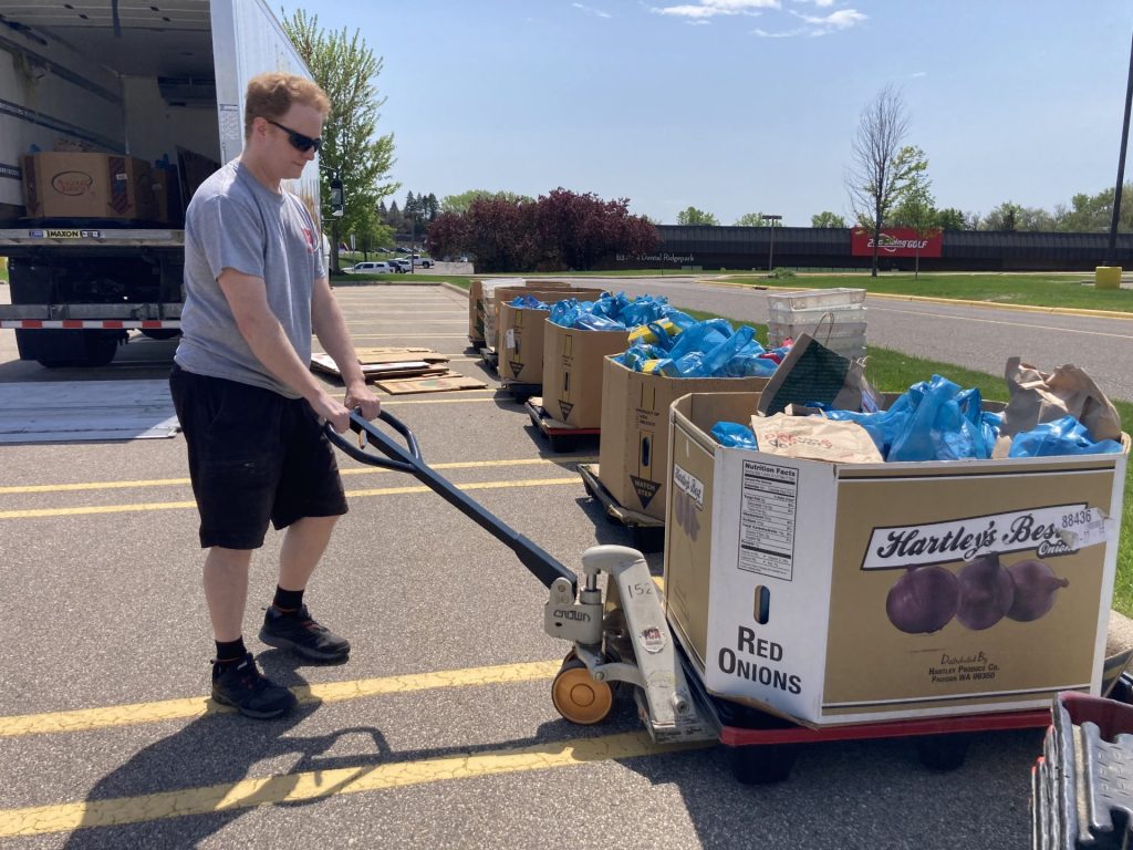 A man wearing sunglasses and shorts uses a pallet jack to move a large box labeled "Red Onions" filled with blue bags during the annual food drive. More boxes and bags line the parking lot, with an American Solutions for Business truck nearby.