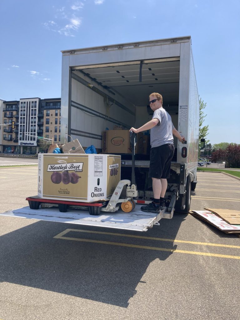 A man stands on the lift gate of a delivery truck, moving a pallet with boxes labeled "Red Onions" for a food drive using a pallet jack in a parking lot on a sunny day. Apartment buildings are visible in the background.