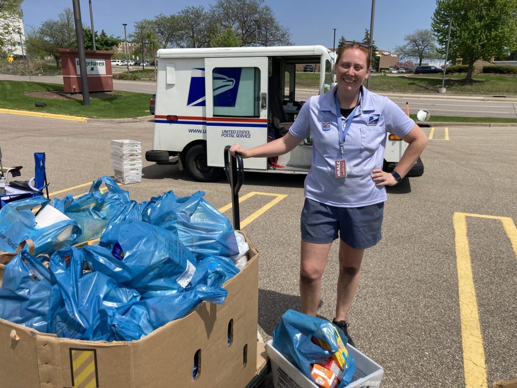 A USPS worker stands next to a bin filled with blue bags of donations, smiling at the camera during the Annual Food Drive. A postal truck is parked behind her in a sunlit parking lot with green trees in the background.