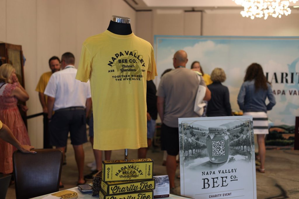 A yellow "Napa Valley Bee Co." t-shirt is displayed on a mannequin at an indoor event. Several people mingle in the background near ASB charity boxes and a sign for the Napa Valley Bee Co. charity event.