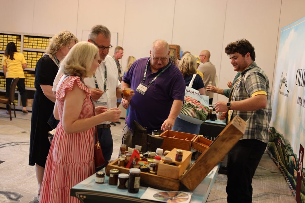 At the ASB Summit in Napa Valley, a group gathers around a display table with jars of jams and condiments. One man points at an item as others look and sample products, enjoying the casual and social atmosphere in California.