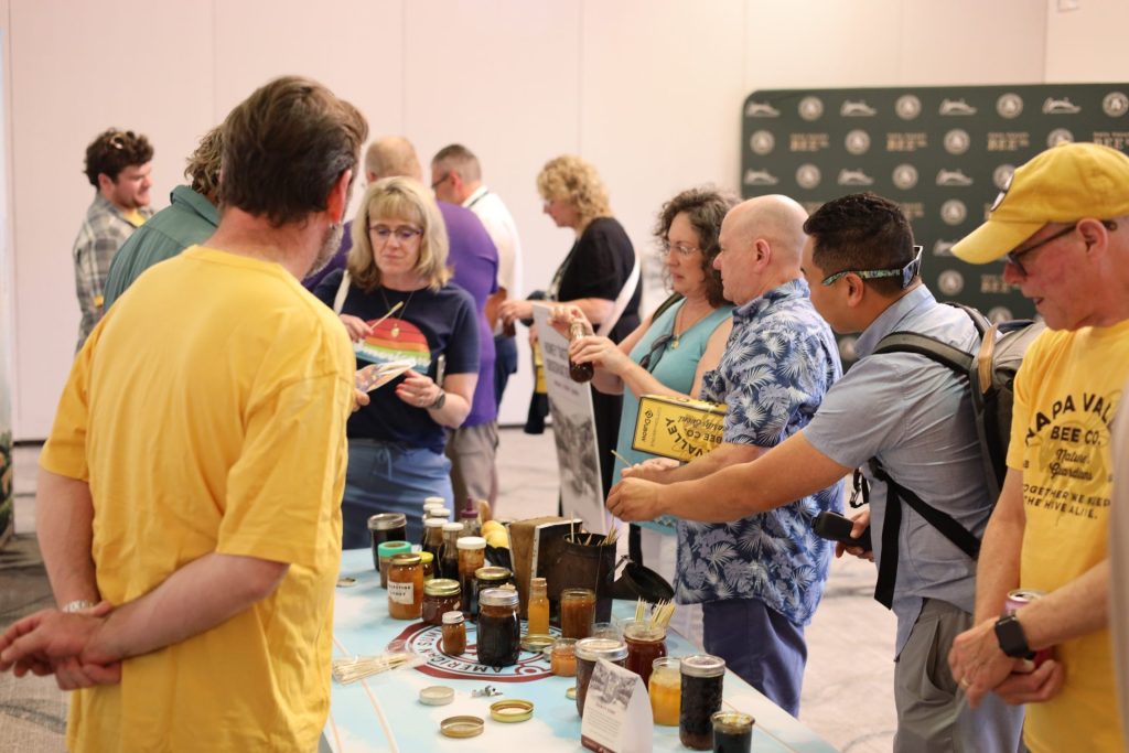People gather around a table displaying Napa Valley Bee honey jars at an indoor fundraising event, examining and sampling the products. Some are reading labels or pamphlets, creating a lively and engaging atmosphere.