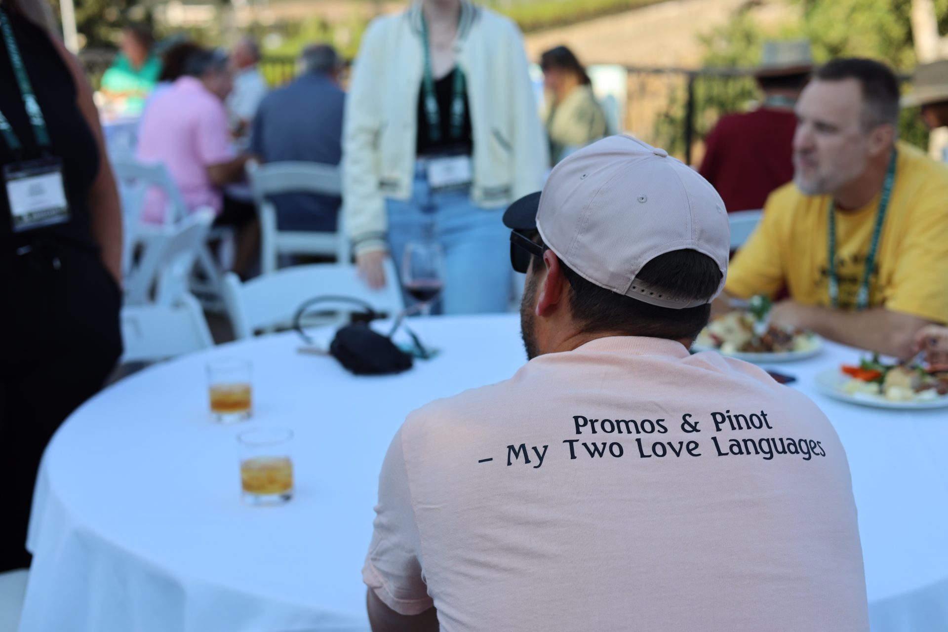 A man sits at an outdoor table in Napa Valley, wearing a hat and a shirt that reads, "Promos & Pinot – My Two Love Languages." Other guests enjoy food and drinks, savoring the relaxed California vibe.