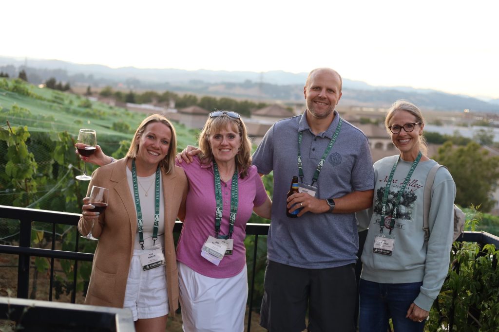 Four adults wearing conference badges smile and pose outdoors by a railing with vineyards and mountains in the background at the ASB Summit in Napa Valley; two hold glasses of wine, one holds a beer.