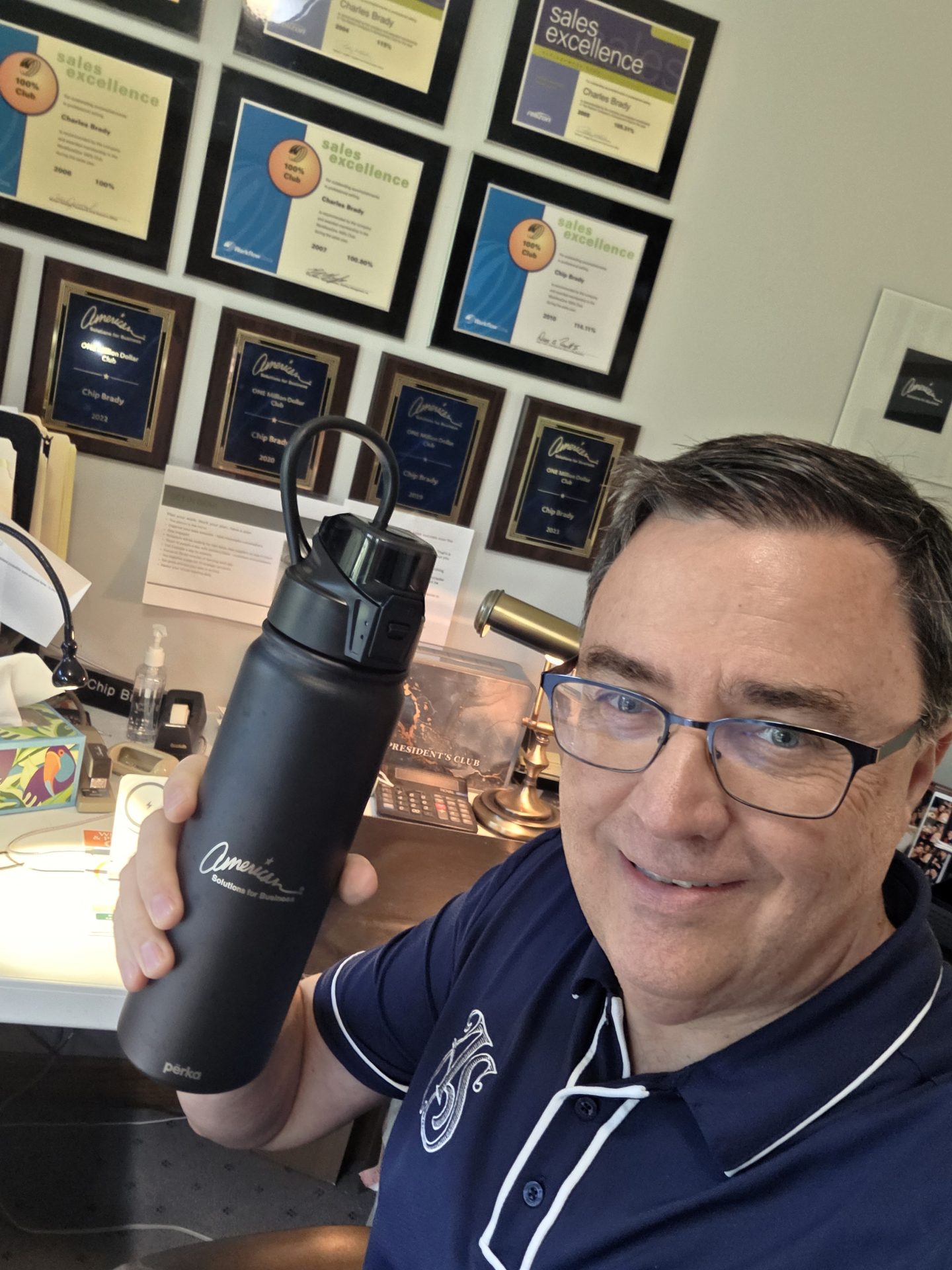Smiling man wearing glasses and a navy collared shirt holds up a black water bottle, highlighting Promotional Products Work Day. Behind him, a wall displays multiple sales excellence plaques above a desk with office items.