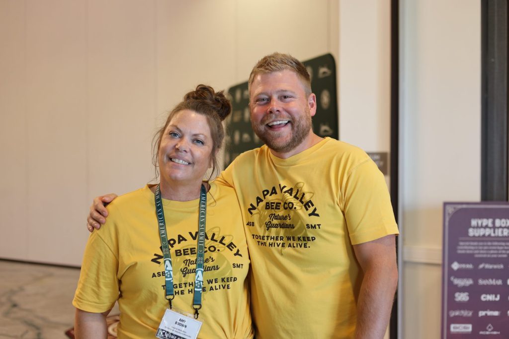 Two smiling people wearing matching yellow "Napa Valley Bee Co." shirts stand indoors at an ASB event. One has a name badge and lanyard, and they stand close together, posing for the photo.