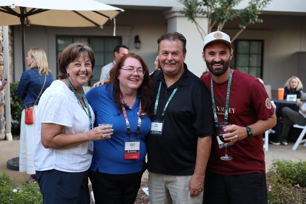 Four adults, three wearing ASB Summit name badges, stand closely together outdoors in Napa Valley, smiling at the camera and holding drinks, with other people and trees visible in the background.