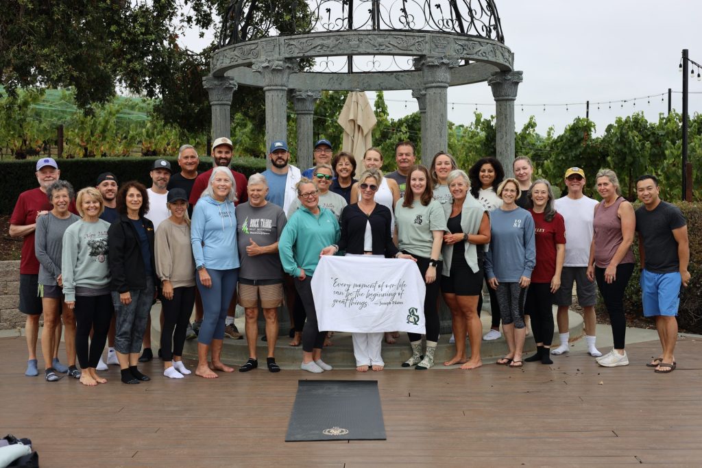 A large group of adults, dressed casually, poses together outdoors on a wooden deck in front of a gazebo at the ASB Summit in Napa Valley. Some hold a white banner, with scenic California grapevines visible in the background.