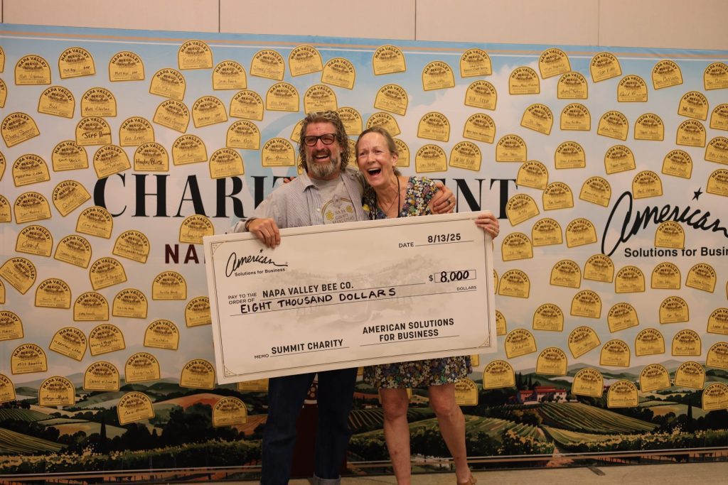 Two smiling people hold a large $8,000 check made out to Napa Valley Bee Co. at a charity event sponsored by ASB. A backdrop covered with bee-themed designs highlights the partnership between Bee Co and ASB.