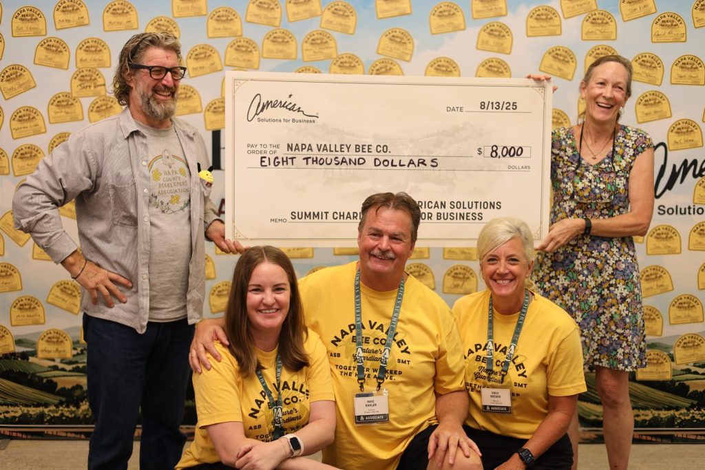 Five smiling people pose with an oversized $8,000 fundraising check made out to Napa Valley Bee Co. Four wear matching yellow shirts with bee-themed text, while two stand and three kneel in front of a bee-themed backdrop.