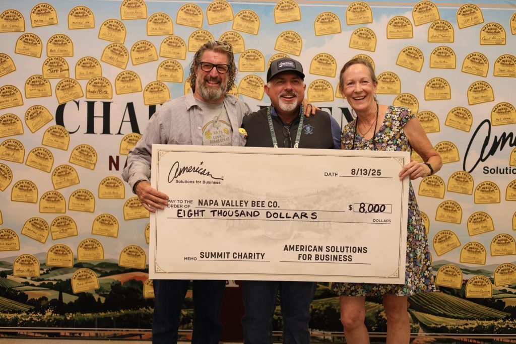 Three smiling people stand together, holding an oversized check for $8,000 made out to Napa Valley Bee from ASB, celebrating a successful fundraising effort against a backdrop of bee-themed plaques.