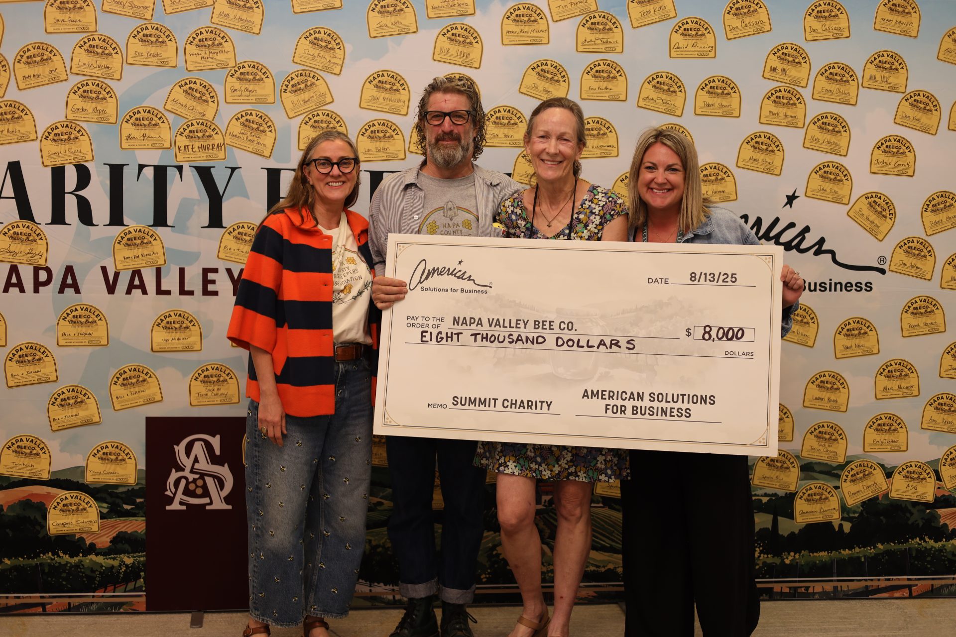 Four smiling people stand together holding a large check for $8,000 made out to Napa Valley Bee Co., celebrating their fundraising success. The background features yellow paper bees and a sign that reads "Charity Fund Napa Valley.