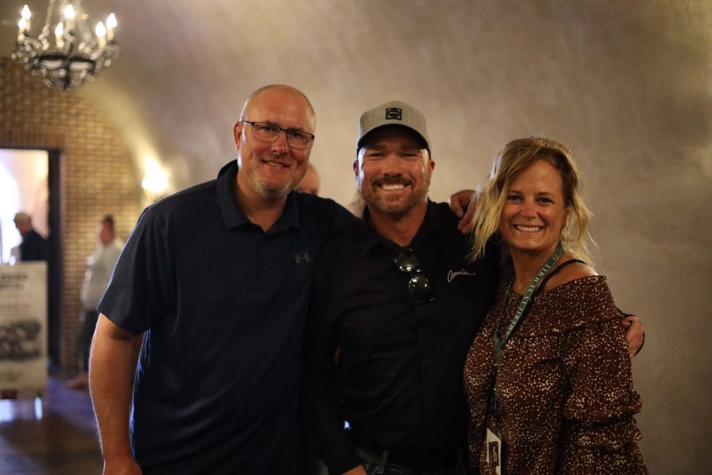 Three adults stand close together indoors, smiling at the camera. Two men are on the left, one wearing glasses and a navy shirt, the other a cap and black shirt. A woman in a brown patterned blouse stands on the right at the ASB Summit in Napa Valley. Warm lighting fills the room.