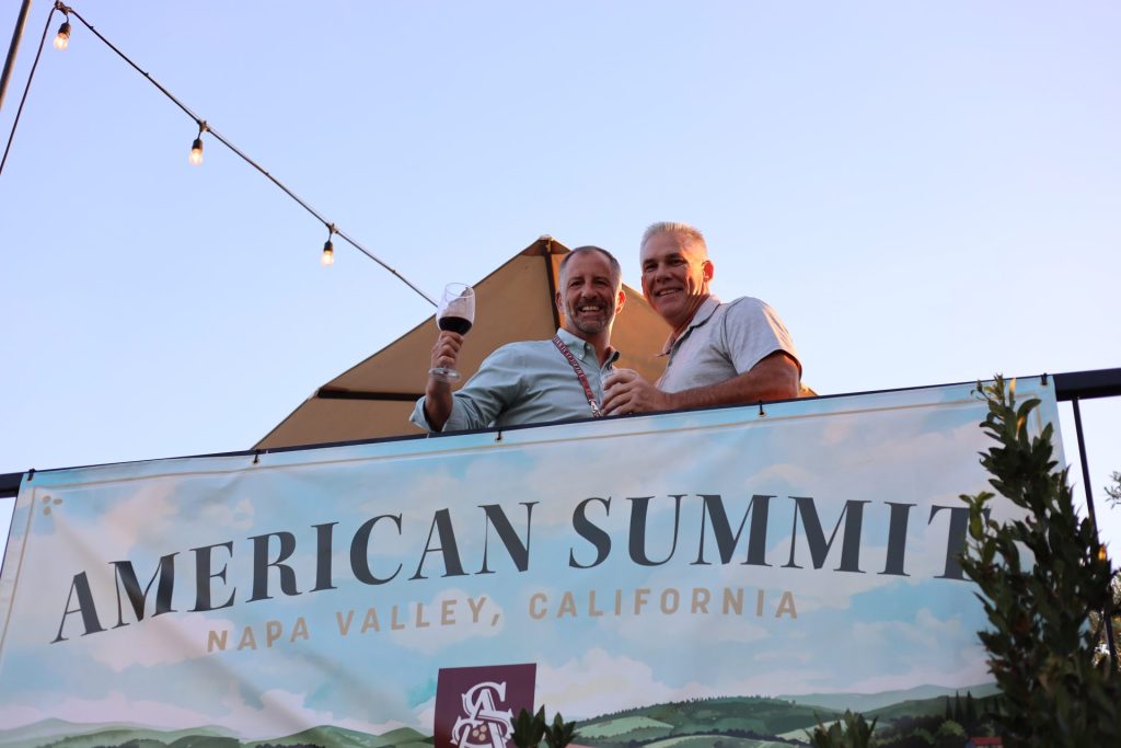 Two smiling men stand behind a large banner reading "ASB Summit, Napa Valley, California," holding drinks outdoors under string lights, with a clear blue sky in the background.
