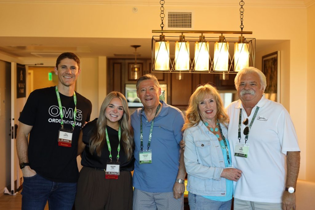 Five people stand indoors under a chandelier, smiling at the camera. Wearing ASB Summit lanyards and casual clothes, they gather in a warmly lit kitchen or dining area, likely in scenic Napa Valley, California.