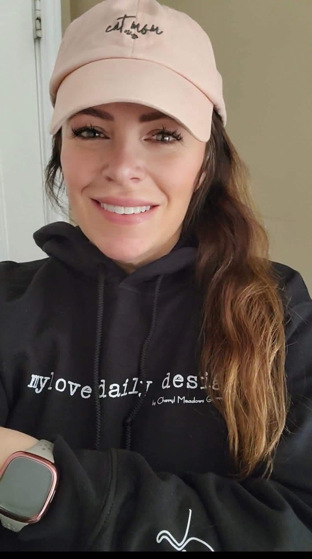 A woman with long brown hair in a ponytail smiles at the camera. She wears a light pink cap, a black hoodie with white text reading "Promotional Products Work," and a smartwatch, standing indoors against a beige wall.