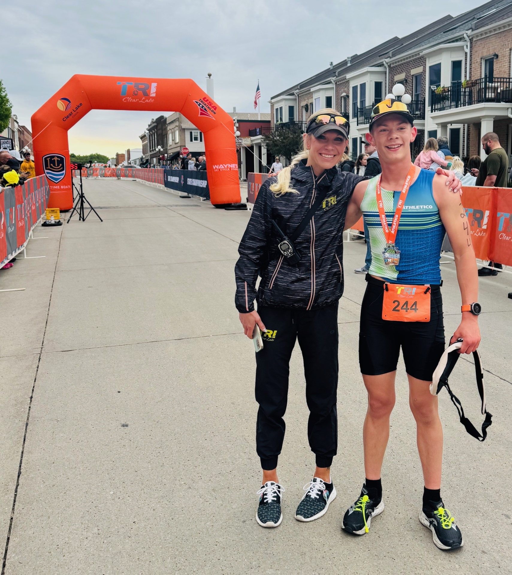 A woman and a young man in athletic gear smile and pose together near a finish line at a triathlon event, with orange race barriers, an inflatable arch, and branded promotional products in the background. The young man wears race bib 244.