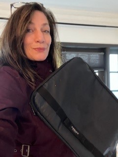A woman with long brown hair, wearing a maroon jacket and glasses on her head, holds a black Targus bag—one of her favorite promotional products—while standing indoors near a window.