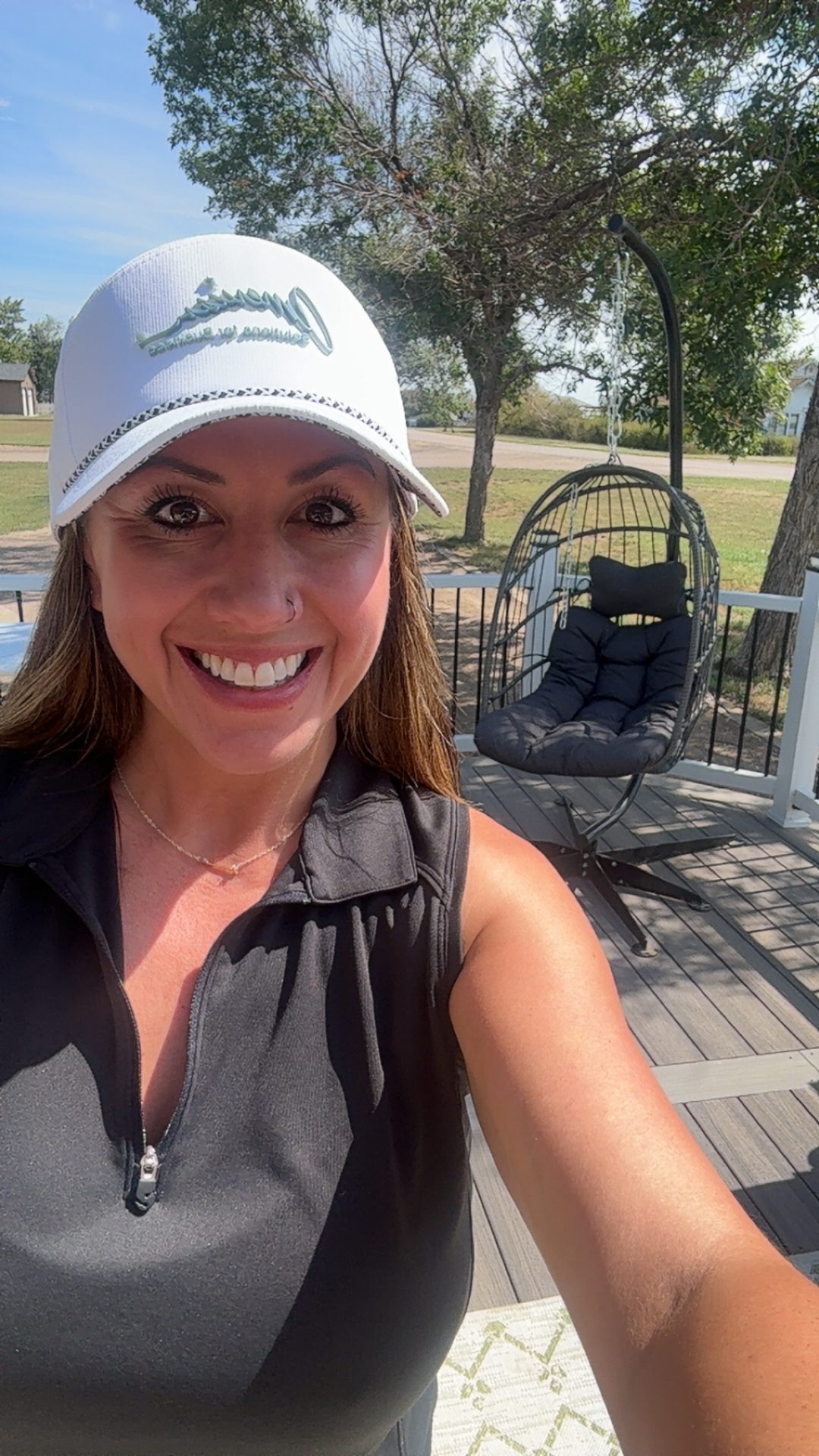 A smiling woman wearing a white cap and black sleeveless top takes a selfie outdoors on a sunny day, showcasing how Promotional Products Work, with a hanging egg chair, wooden deck, trees, and grassy area in the background.