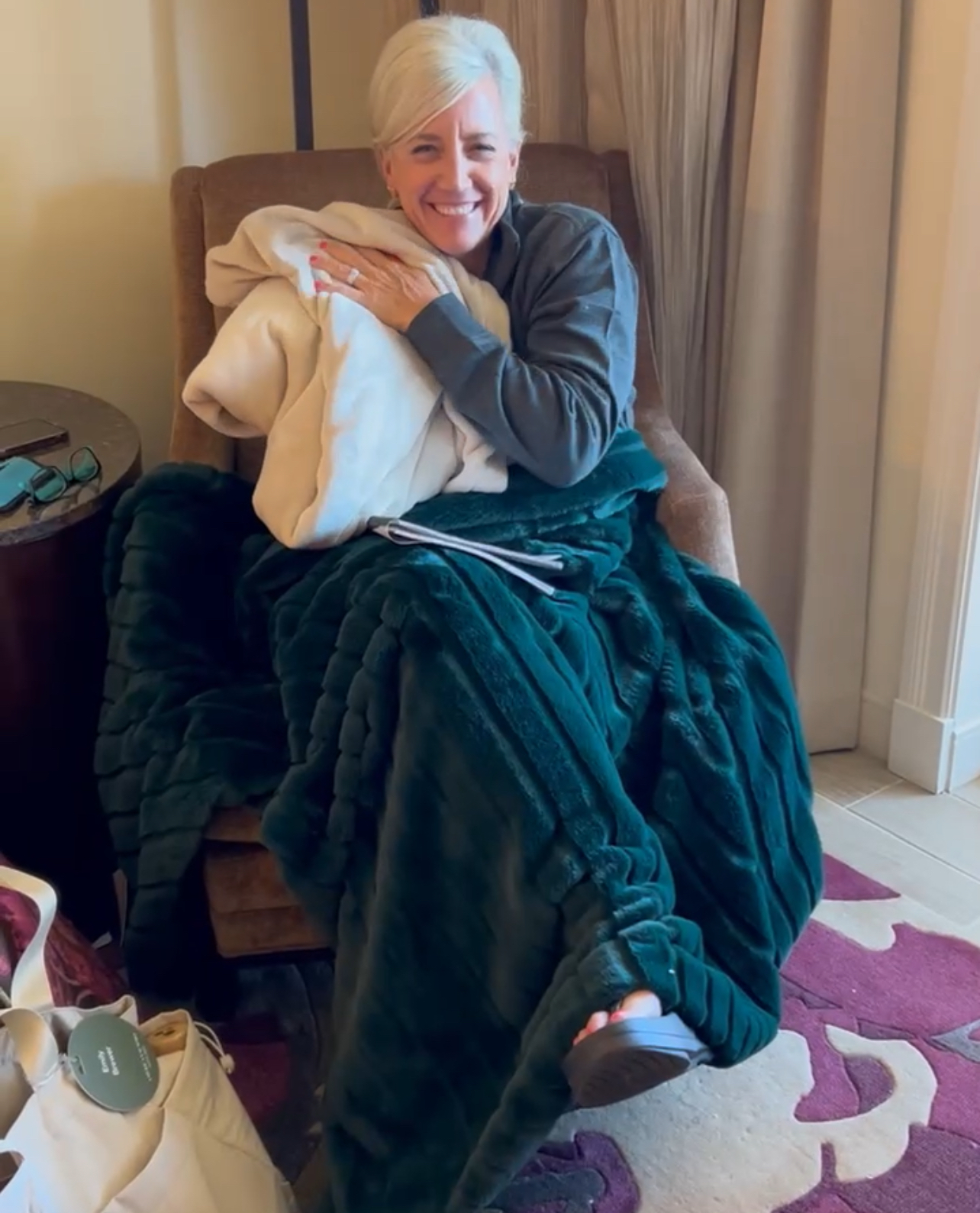 A woman with short gray hair sits smiling in an armchair, wrapped in a dark green blanket and hugging a cream-colored blanket—looking cozy and relaxed, with glasses, a tablet nearby, and enjoying her favorite Promotional Products.