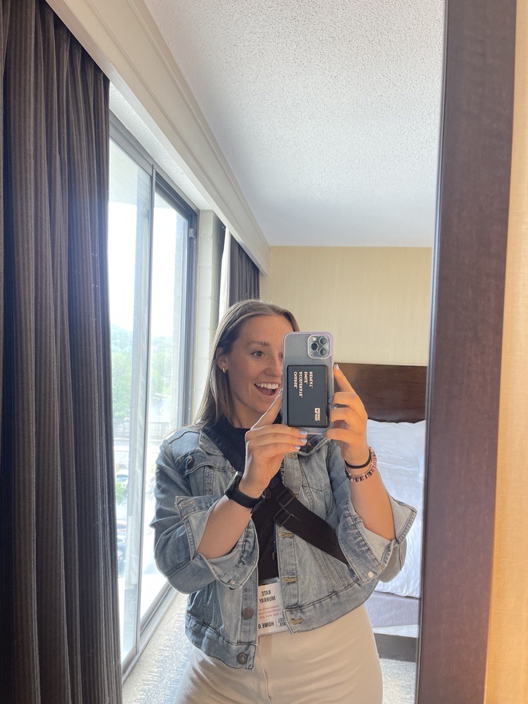 A woman smiles while taking a mirror selfie in a bright hotel room on August 28th 2025. She wears a light denim jacket, black crossbody bag, and name badge promoting Promotional Products Work, with sunlight streaming through large windows.