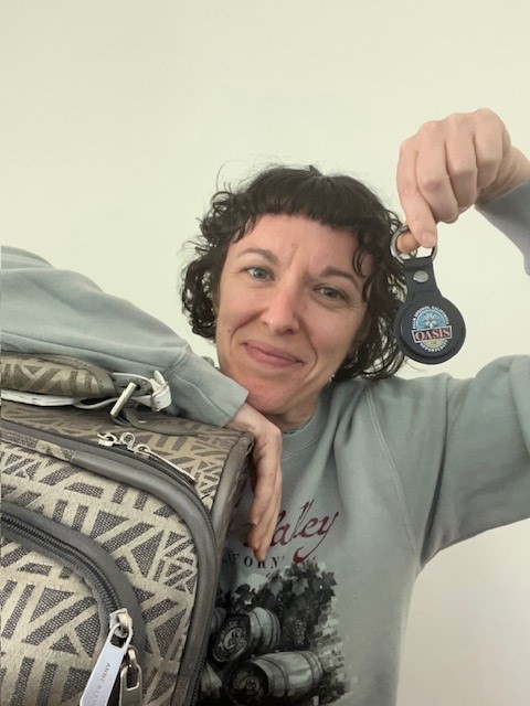 A smiling person with short curly hair holds up a promotional products-themed keychain and leans on a patterned suitcase against a plain background, celebrating the impact of Promotional Products Day.