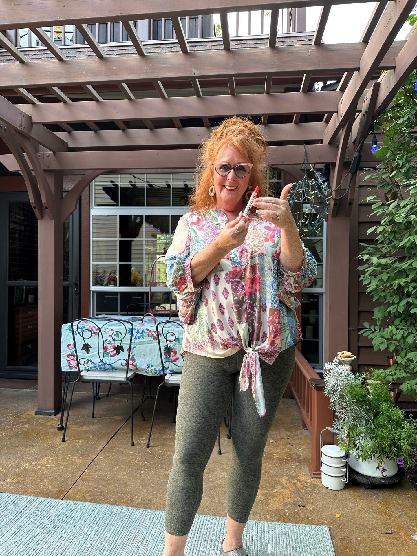 A smiling woman with long red hair, glasses, and earrings stands on a patio under a wooden pergola. Wearing a floral blouse and olive leggings, she holds a vape pen, gesturing playfully—celebrating Promotional Products Day among outdoor decor.