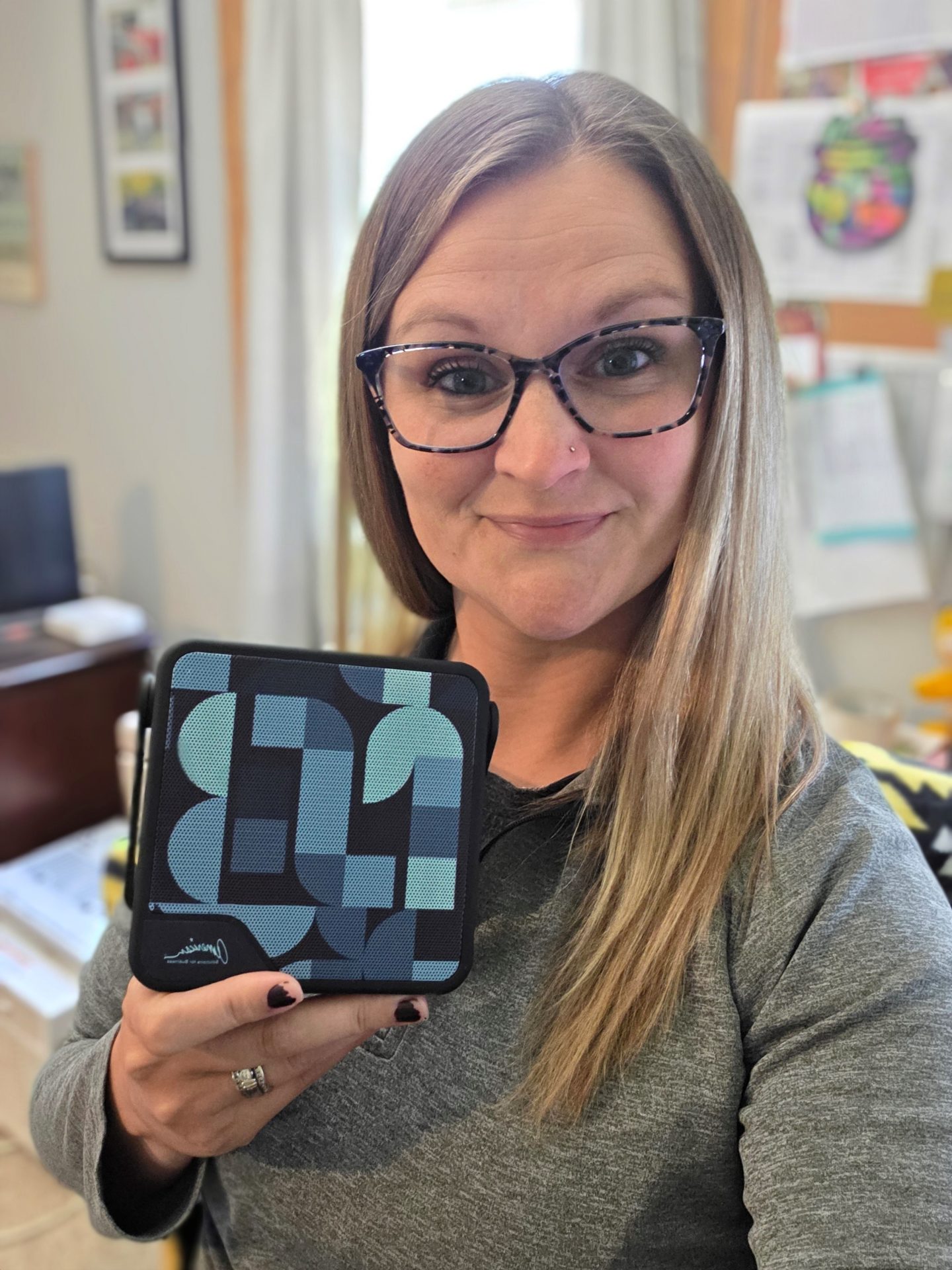 A woman with long blonde hair and glasses smiles while holding up a small, patterned square case in a bright home office, celebrating Promotional Products Work Day on August 28th, 2025.