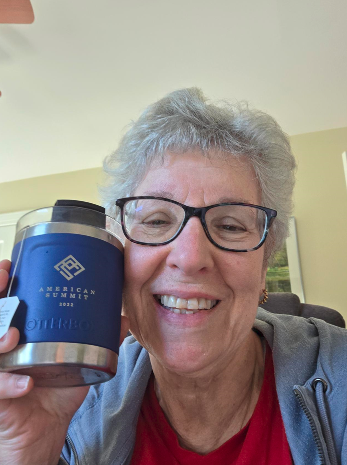 An older woman with short gray hair and glasses smiles at the camera while holding a navy blue “American Summit 2023” promotional product tumbler. She wears a red shirt and gray sweatshirt. Indoor background with soft lighting.