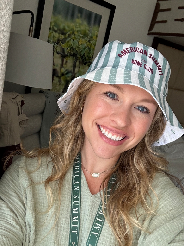 A smiling woman wearing a striped bucket hat that says "American Summit Wine Club" and a lanyard stands indoors with art and a jacket in the background, celebrating Promotional Products Work with custom gear on August 28th, 2025.
