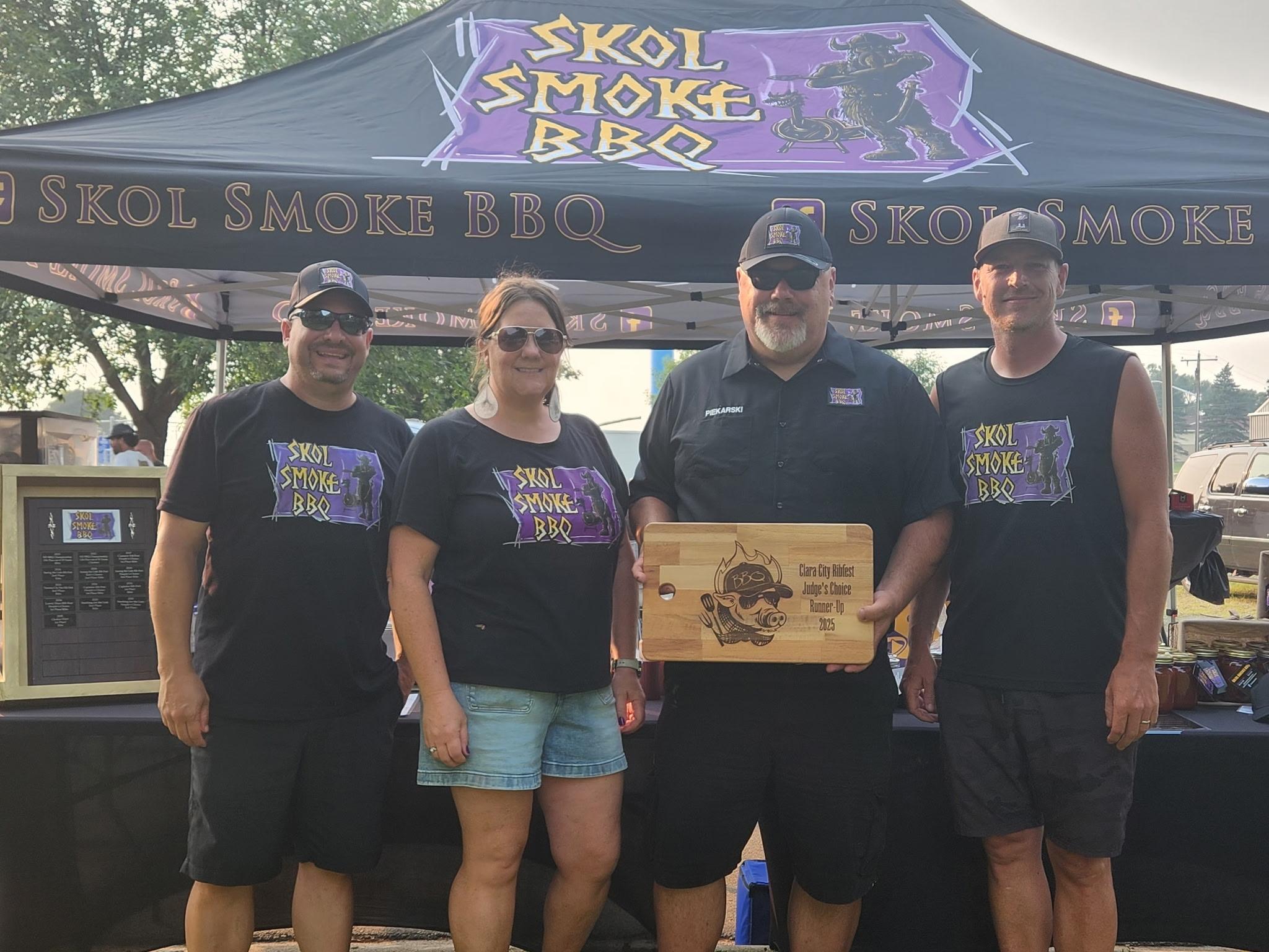 Four people in matching "Skol Smoke BBQ" shirts celebrate Promotional Products Work Day in front of a BBQ tent. Two men wear shorts and caps; one holds a wooden award plaque. A woman in sunglasses smiles, with BBQ equipment behind them.