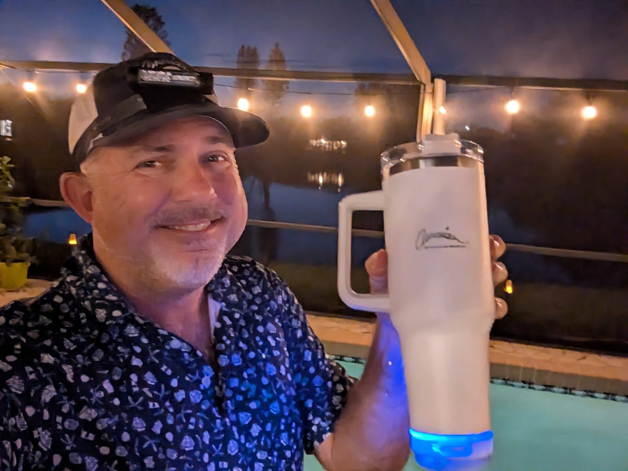 A smiling man in a patterned shirt holds a large white tumbler with a blue LED base by a pool at night, showcasing how Promotional Products can light up any evening. String lights and water shimmer in the background.