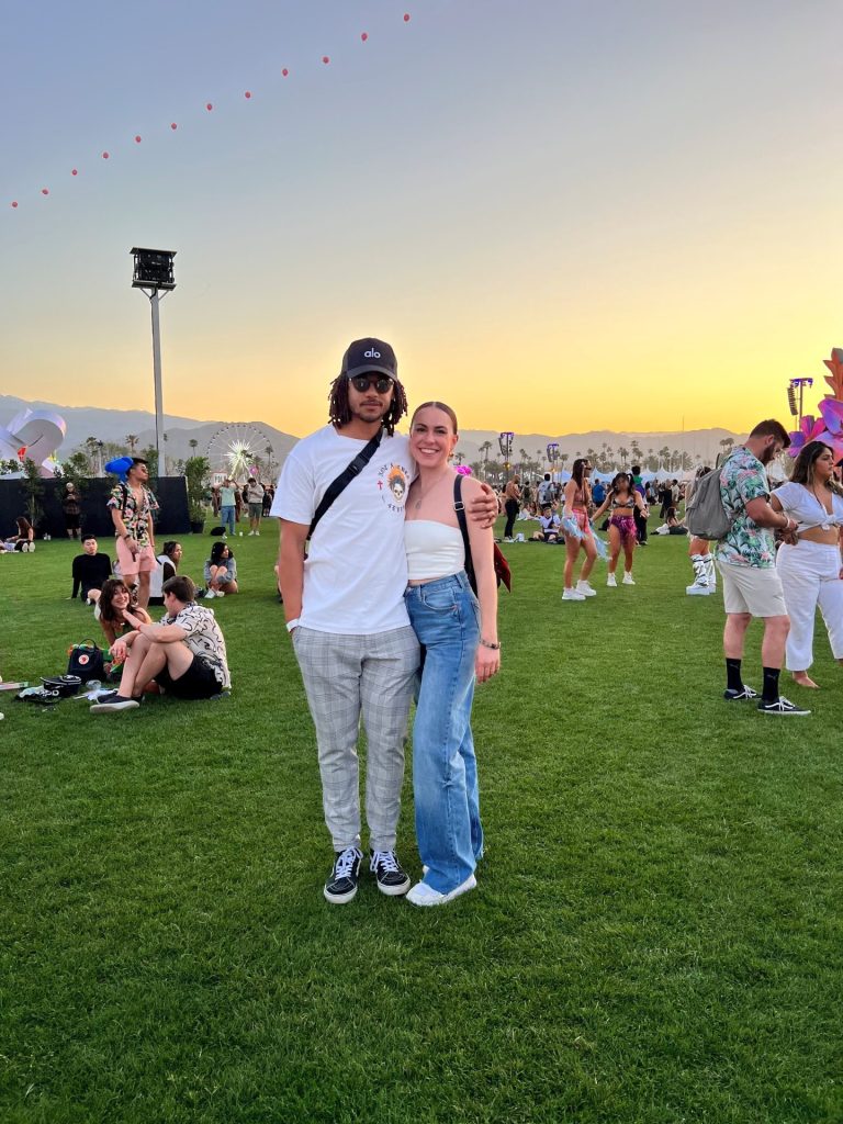 Two people pose together on a grassy field at an outdoor festival during sunset, with Megan Spire among the attendees. The sky is clear, and festival decorations and palm trees are visible in the background.